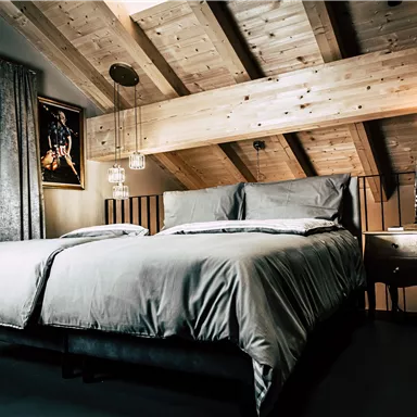 A modern bedroom with a large double bed and wooden beam ceiling. The walls are light, and there are large windows that let in natural light.