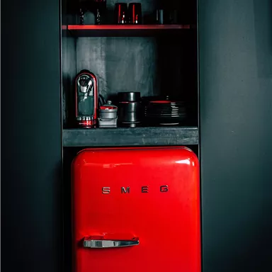 A red refrigerator stands in a modern cabinet. Above it are stylish dishes and a coffee machine.