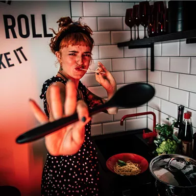A cheerful woman is cooking in a modern kitchen. She is holding a spoon in her hand and seems to be having fun cooking.