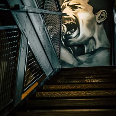 A staircase with dark wood and a metallic railing. On the wall hangs an impressive mural of a screaming man.