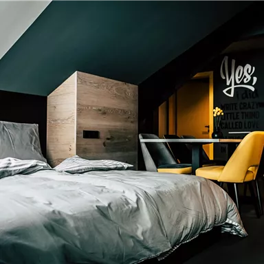 A modern bedroom with a large bed and a stylish table. The walls are dark, and there are yellow chairs that create a contrast.