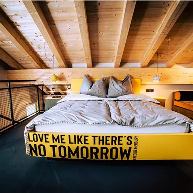 A modern bedroom with a large bed and wooden ceiling. The bed frame has the inscription "Love me like there’s no tomorrow."
