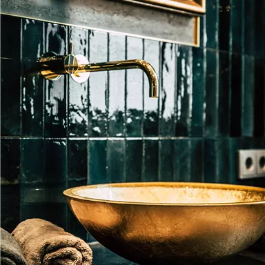 An elegant gold sink on a dark table. In the background, green tiles and a golden frame can be seen.