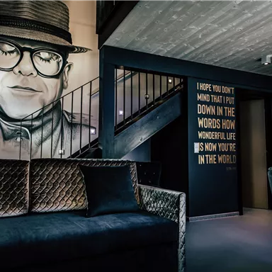 A stylishly furnished room with a gray sofa and a large mural of a man with glasses. On the opposite wall, there is an inspiring quote in large letters.