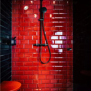 A modern shower with red tiles and a black rain shower. A small red stool is next to the shower.