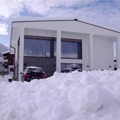 A modern house in a snowy landscape. The snow piles up in front of the building and the sky is partly cloudy.