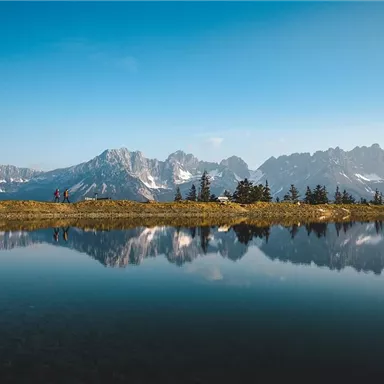 A tranquil mountain lake with clear reflecting water. In the background, impressive mountains and a blue sky can be seen.