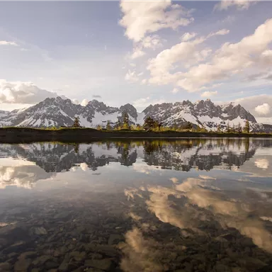 A picturesque mountain landscape with snow-covered peaks is reflected in the clear water of a lake. The sky is adorned with gentle clouds, and the scene is peaceful.