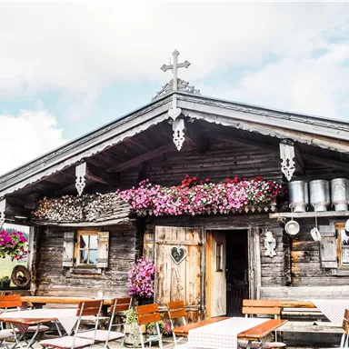 Ein gemütliches Holzhaus mit bunten Blumen und bayerischer Dekoration. Vor dem Haus stehen Tische und Stühle in einem schönen Außenbereich.
