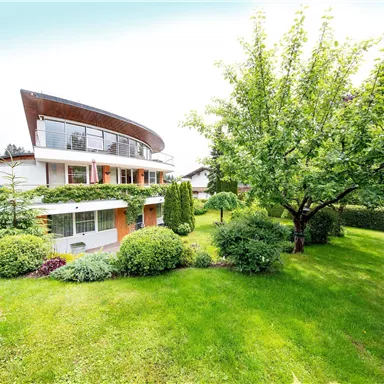 A modern house with a curved roofline surrounded by a well-maintained garden. Green meadows and trees give the scene a tranquil atmosphere.