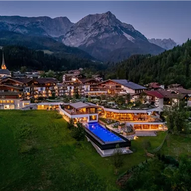 Eine malerische Landschaft mit einem Hotelkomplex, umgeben von Bergen und Wäldern. Der Außenbereich verfügt über einen Pool, der von Lichtern beleuchtet wird.