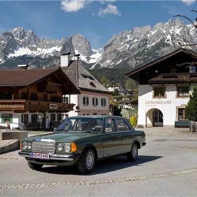 An old Mercedes-Benz drives through a picturesque village with traditional architecture. In the background, majestic mountains rise under a clear sky.