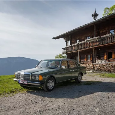 An older car is parked in front of a traditional wooden house on a hill. In the background, a picturesque mountain landscape stretches out.