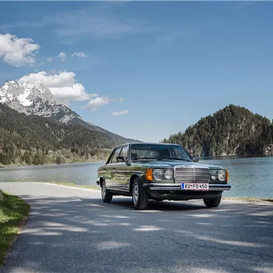 A classic car is parked on a picturesque road along a clear lake. In the background, mountains and a blue sky can be seen.