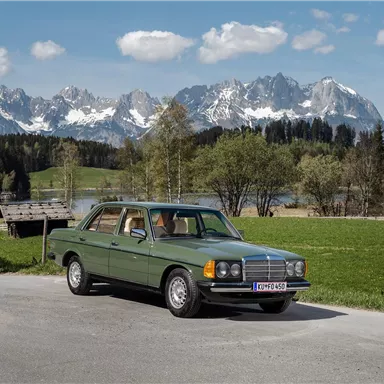 A classic car is standing on a road in front of a picturesque mountain landscape. In the background, snow-capped mountains and green meadows can be seen.