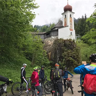 A group of cyclists stands on a path, surrounded by lush greenery. In the background, a small church with a distinctive dome can be seen.