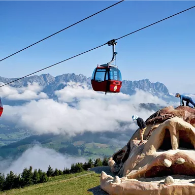Eine Seilbahn schwebt über eine beeindruckende Berglandschaft. Auf einem großen Kunstwerk in Form eines Gesichts klettern zwei Personen.