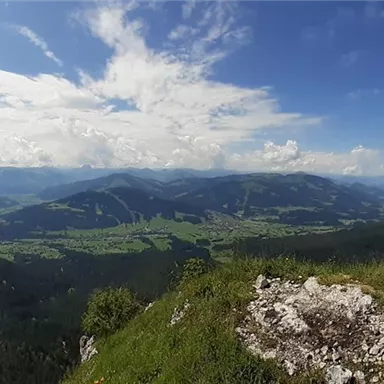An impressive mountain view with green meadows and imposing peaks. The sky is blue and slightly cloudy.
