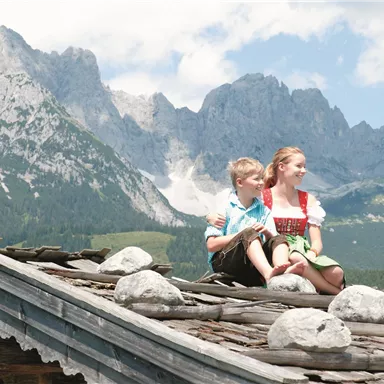 Two children are sitting on a roof and enjoying the view. In the background, majestic mountains and a blue sky can be seen.
