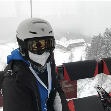 A person in ski gear is sitting in a gondola on the way to the ski resort. In the background, snow-covered trees and a house can be seen.