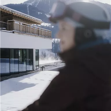 A snowy outdoor area with a modern building in the background. In the foreground, there is a person with ski equipment and ski goggles.
