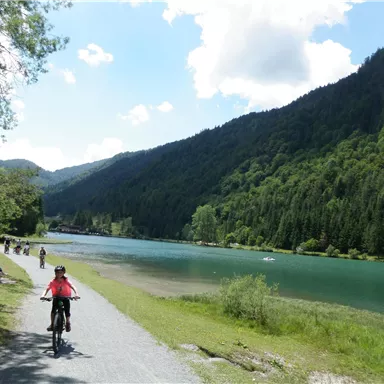 A cyclist rides along a path beside a clear, blue lake. In the background, green mountains rise under a bright blue sky.