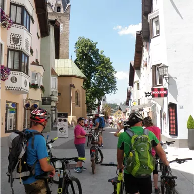 A group of cyclists stands on a picturesque street with historical buildings. In the background, a church tower and blooming trees can be seen.