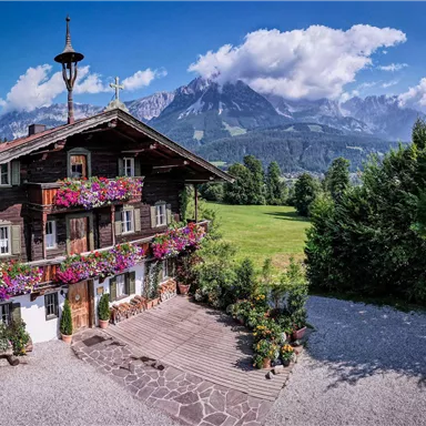 Ein charmantes Holzhaus mit bunten Blumen auf den Fensterbänken steht vor einer beeindruckenden Bergkulisse. Der Himmel ist klar und die Natur um das Haus ist grün und einladend.