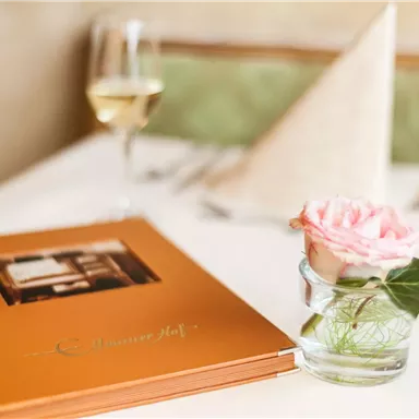 An elegant table with a menu and a rose in a glass vase. In the background, there is a glass of wine and a folded napkin on the table.
