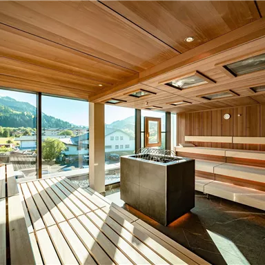 A modern sauna interior with wooden shelves and large windows. The view extends over a picturesque landscape with mountains.