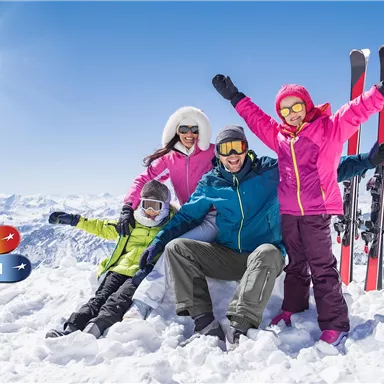 A happy family stands in the snow and poses for a photo. In the background, snow-covered mountains and skis are visible.