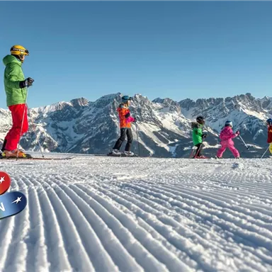A group of skiers on a snow-covered slope with impressive mountains in the background. The sky is blue and the scene shows activities in the snow.