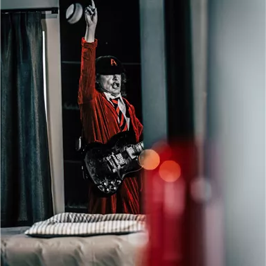 A musician in a red outfit is playing guitar in a modern room. In the background, a bed with striped pillows is visible.