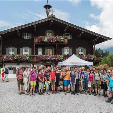 A large group of people stands in front of a traditional, flower-decorated chalet in the mountains. In the background, green hills and a cloudy sky are visible.