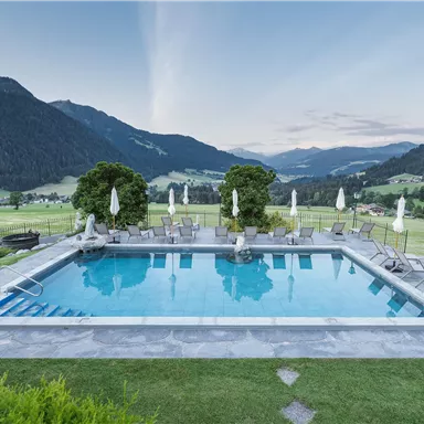 A beautiful pool with sun loungers, surrounded by green meadows and mountains. The sky is clear and shows soft colors of the sunset.