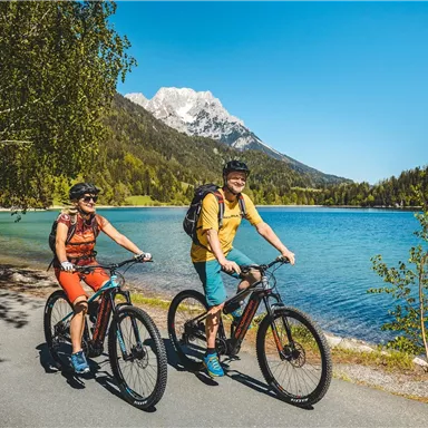 Two cyclists ride along a clear lake in a picturesque landscape. In the background, mountains and a bright blue sky can be seen.