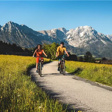 Two cyclists are riding on a path through a blooming landscape. In the background, impressive mountains can be seen.