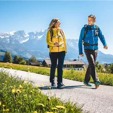 A couple is hiking on a picturesque path in the mountains. The sunny landscape is surrounded by green meadows and snow-covered peaks.