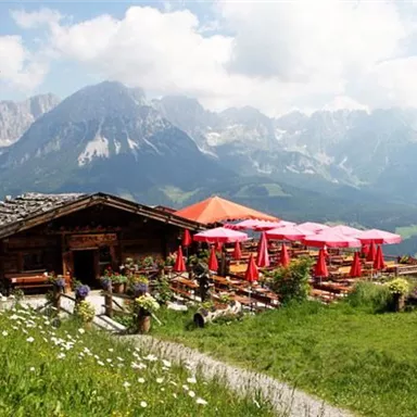 A picturesque mountain cabin with a terrace and red sunshades. In the background, impressive mountains and a blue sky can be seen.