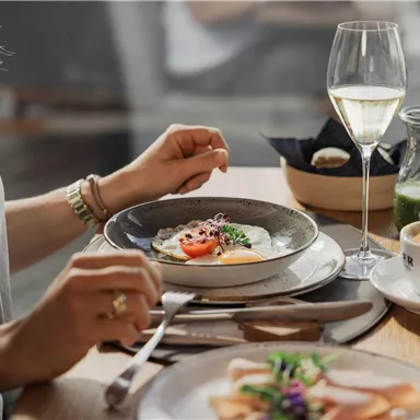 An elegantly set table with various dishes and a glass of wine. A hand holds cutlery, while a cup of coffee and fresh flowers are visible in the background.