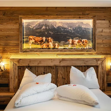 A cozy bedroom with a wooden bed and two large pillows. On the wall hangs a picture of cows in a mountain landscape.