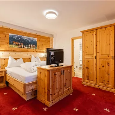 A cozy bedroom with wooden decor and a red carpet. The room features a king-size bed, a television, and a large wardrobe.
