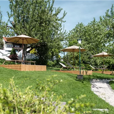A green garden area with sun umbrellas and loungers. The space is surrounded by trees and offers an inviting, quiet place to relax.