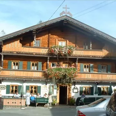 A traditional Alpine house with a wooden-clad facade and colorful flowers. The entrance has an inviting atmosphere with outdoor tables.