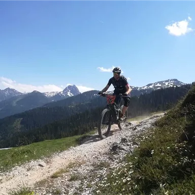 A cyclist rides on a gravel path through the mountains. The sky is clear and the landscape is green and mountainous.