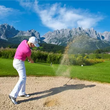 A golfer is hitting from a bunker on a beautiful golf course with impressive mountains in the background. The sky is clear and blue, the landscape is green and inviting.