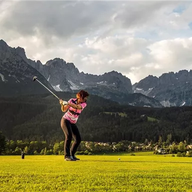 A female golfer is hitting a shot in a picturesque landscape with mountains in the background. The green grass and the cloudy sky create an impressive backdrop.
