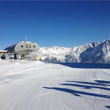 A snow-covered mountain landscape with a lift station in the foreground. In the background, majestic mountains can be seen under a clear blue sky.