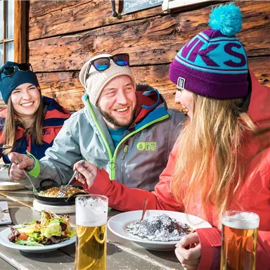 A group of three friends is sitting relaxed at a table in a mountain lodge. They are enjoying food and drinks in a cozy atmosphere.