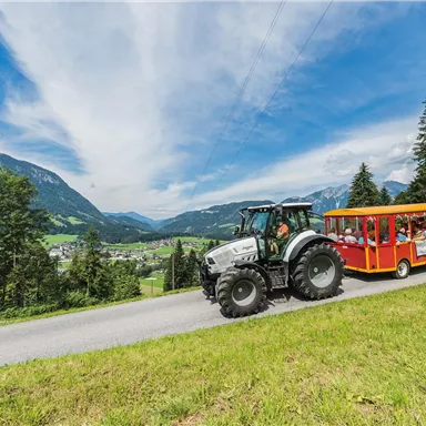 Ein Traktor zieht einen bunten Wagen mit Passagieren auf einer kurvenreichen Straße. Im Hintergrund sind grüne Hügel und ein klarer Himmel zu sehen.